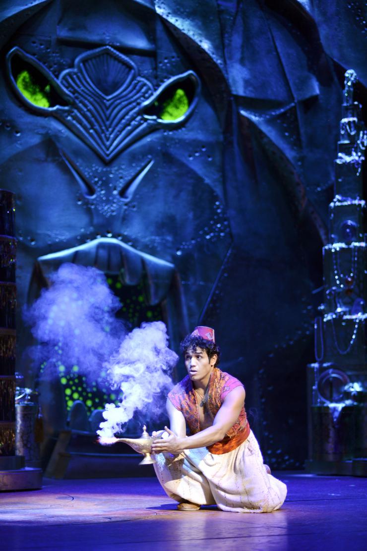 An actor on stage squats while rubbing the side of a genie lamp.