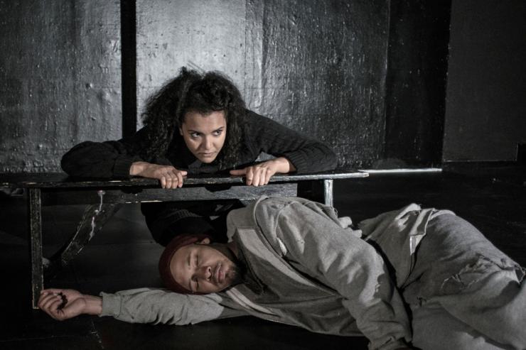 One actor sleeping beneath a bench while another leans on it