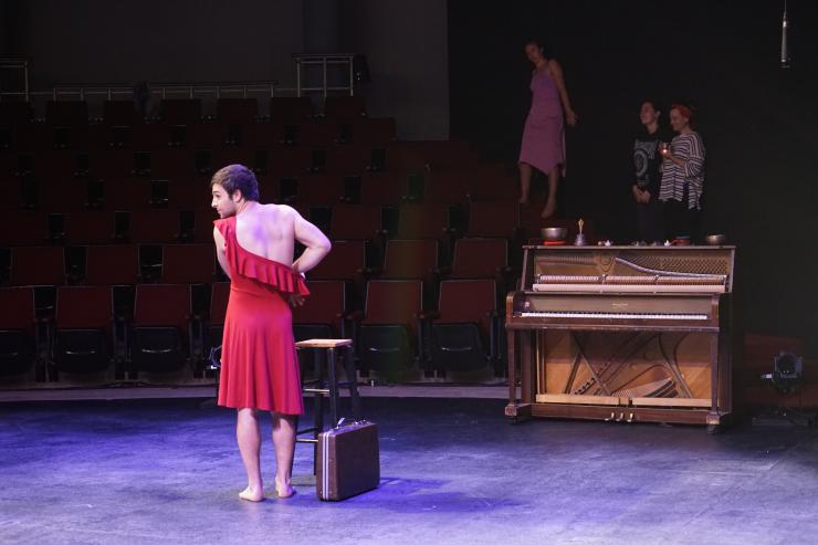 A man wears a red dress on stage next to a piano