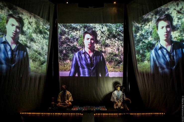 Two actor sit on stage under the projected videos of a man 