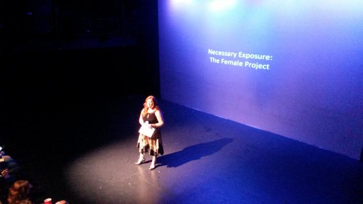 Woman on stage in front of a projection that reads: "Necessary Exposure: the Female Project."