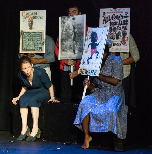 A white performer crouches and leans forward while four other performers hold signs.