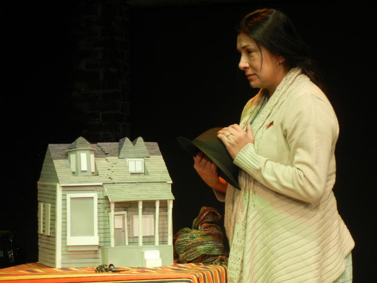 A performer holding a hat stares off left while a model house sits on the table next to them.