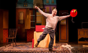A man on stage poses with his arms outstretched as he holds a balloon and makes a comical face.