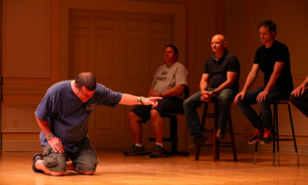 an actor kneeling onstage before four seated actors