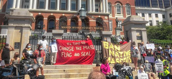 A group of people in front of the Massachusetts State House holding a banner that reads "It's time to change the mass flag and seal" and "ban the native mascots".