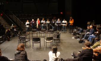 a large group sitting in a circle watching a staged reading