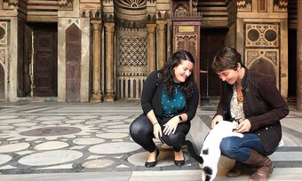 two people kneel in front of ornate architecture, petting a black and white cat