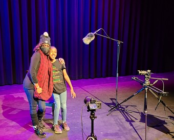 Image of an adult and child standing on a stage with recording equipment.