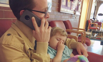 A man is sitting holding a phone up to his ear while a toddler sleeps on him. The toddler is holding a rainbow colored stuffed animal.