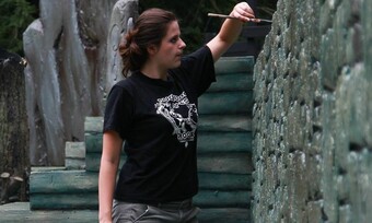 Ashley Malafronte in a black shirt and kakis painting on a stone wall.