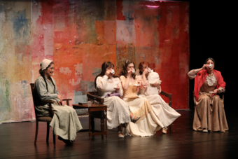 Five women in dresses drinking tea on stage.