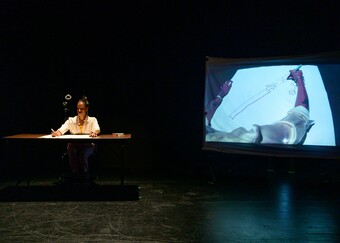 A performer sits at a desk and draws while a screen to the right broadcasts a video of the drawing.