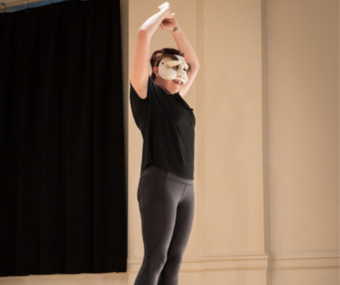 A performer wearing black and a mask standing on top of a box with arms in the air.