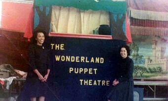 two women in 1950s clothing stand in front of a puppet theater proscenium frame with the wonderland puppet theater written on its front.