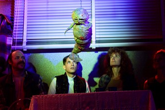 Four performers seated at a table look upward at a worm prop.