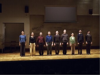 Nine performers stand side by side on stage and look outward to the audience.