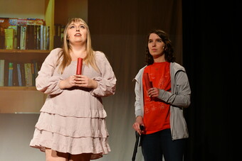 Two actors stand onstange, one in a pink dress and the other in a t-shirt using a cane.