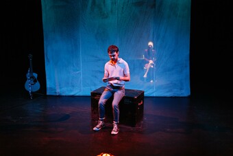 A man sits on stage on a black box in front of a blue scrim.