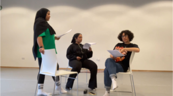 Three young woman sit in chairs reading scripts as a part of a workshop.