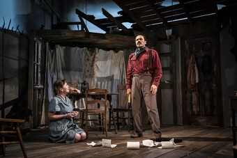 Two actors stand on stage in front of a set made of damaged wood.