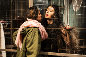 Two women stand face to face on either side of a chain link fence. 