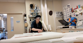 A woman in a black hat cuts fabric in a costume shop.