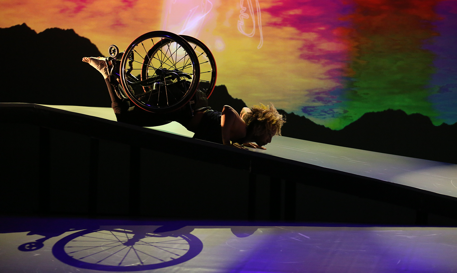 As the sky transitions from gold to amber to red, green and blue, Alice Sheppard, a light-skinned multiracial Black woman, slides in a spider position on her stomach down the shiny ramp. Her bent elbows, her wheelchair and the bottoms of her bare feet point upward as she stares down the Ramp. The shadow of her wheelchair is visible beneath her short curly hair glows in the light.