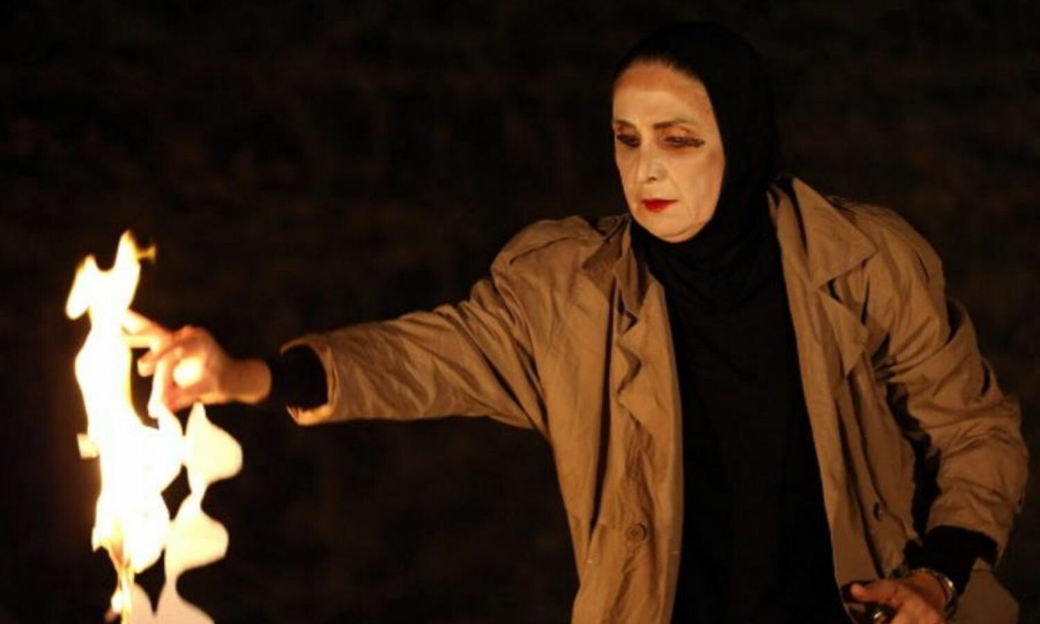A woman in a black hijab and tan trenchcoat burns a piece of paper cut out in a wavy design.