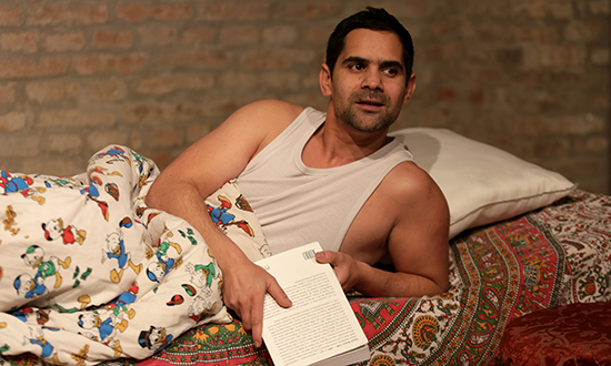 a man in an undershirt sitting up in bed holding a book.