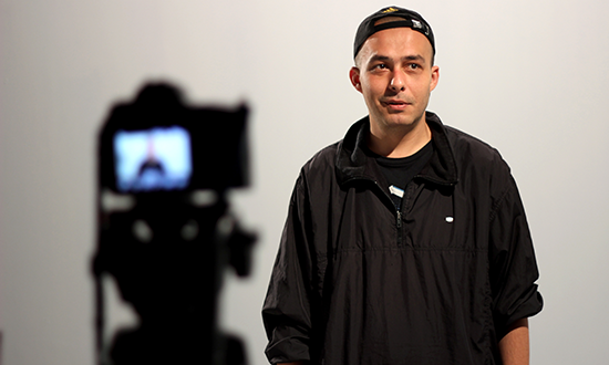 A man in backwards baseball cap stands in front a camera tripod.