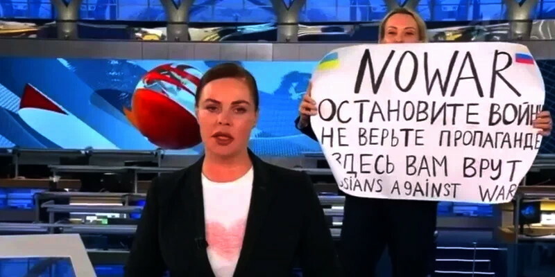 A woman with a sign stands behind a news anchor.
