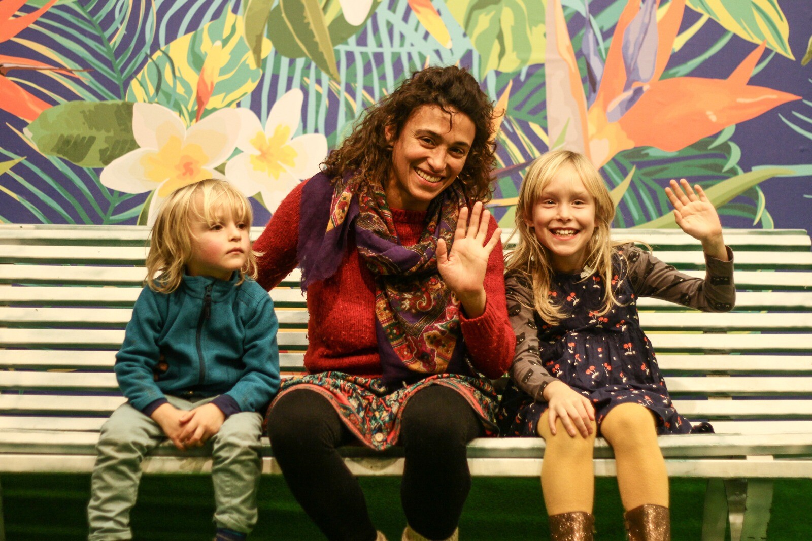 One adult and two kids sitting on a bench and waving at the camera.