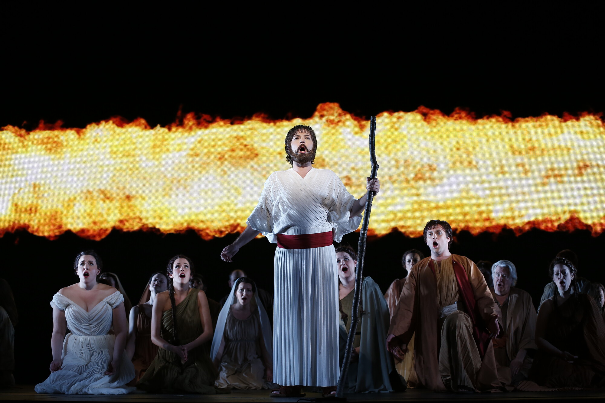 A performer holding a staff sings in front of many other kneeling performers, with a projected image of fire behind them.