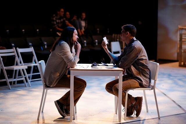 Two actors sit across from each other at a small square table during a show.