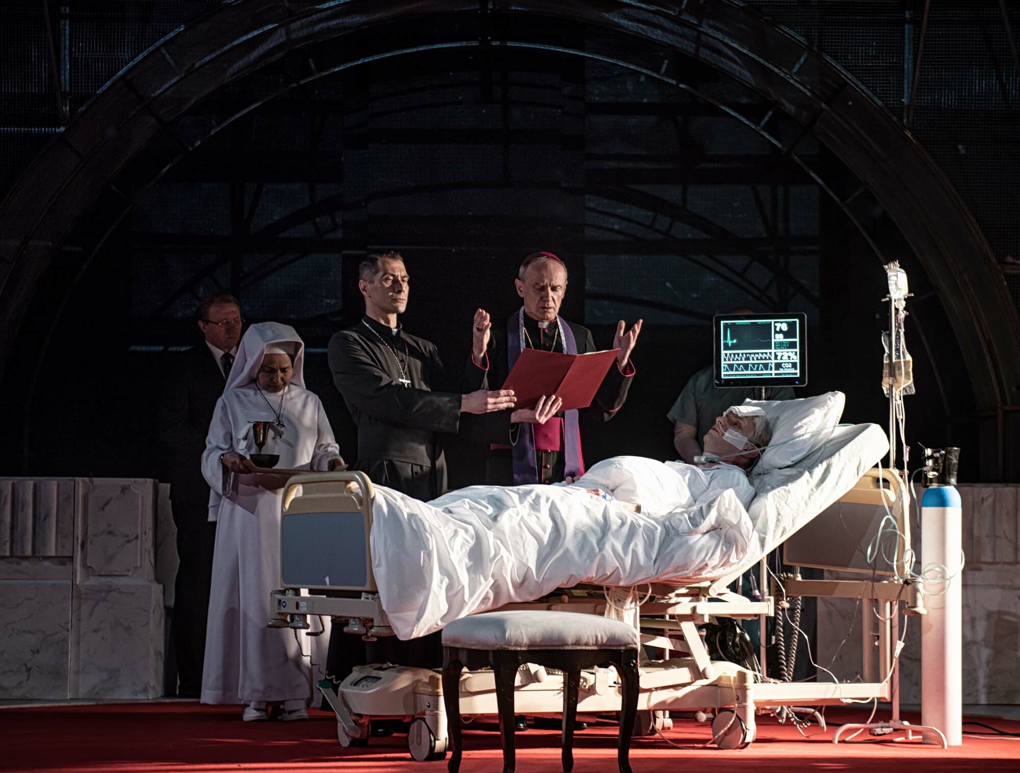 Two actors in costume as priests stand and pray over another actor portraying the bedridden Pope John Paul II, while two other performers look on from upstage.