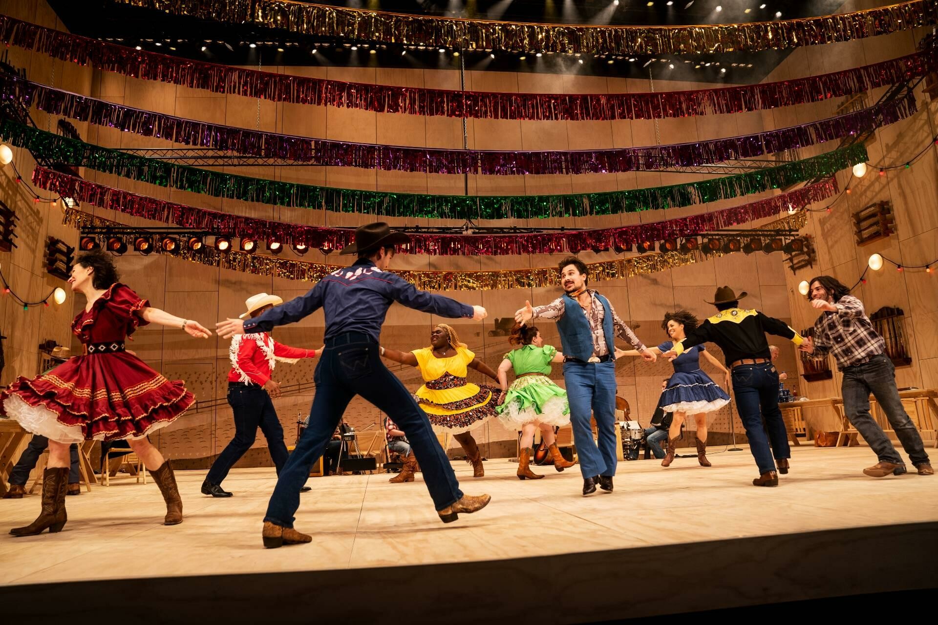 The cast of Oklahoma dancing on a wide well lit stage.