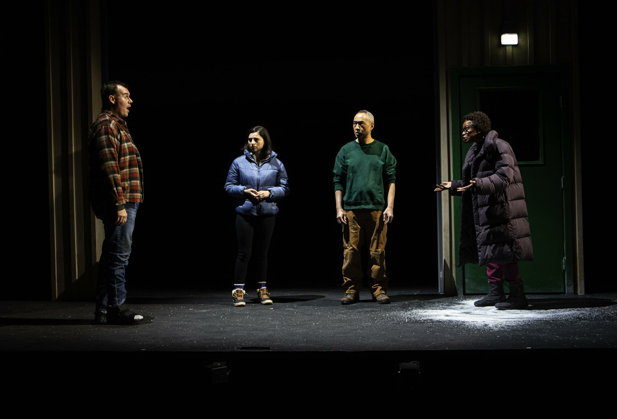 Four performers stand on a dimly lit stage and speak to each other.