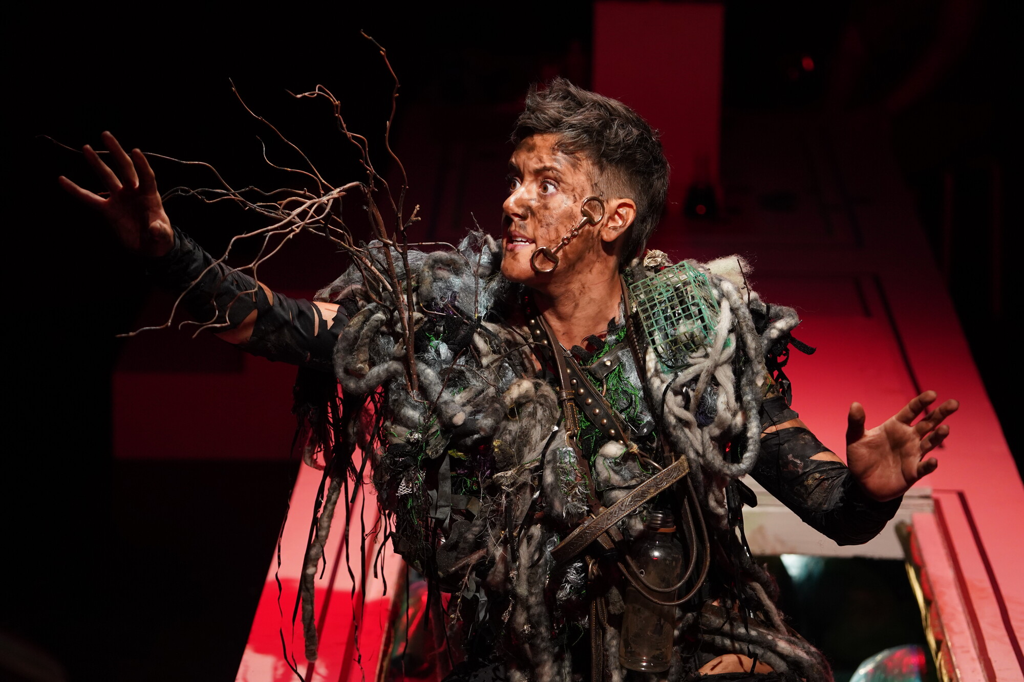 A performer in a costume resembling a combination of trash and natural debris reaches out to the audience with a fierce expression.