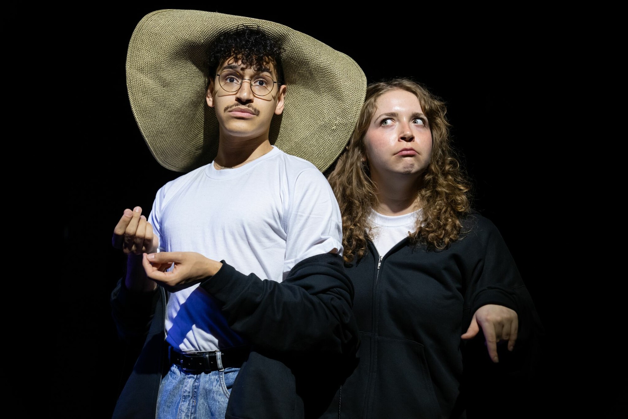 A performer in a large-brimmed floppy straw hat and a performer with a black zip-up hoodie stand onstage with quizzical facial expressions.