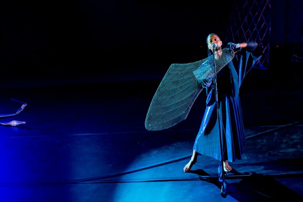 A woman holding a large fan sings into a microphone on stage.