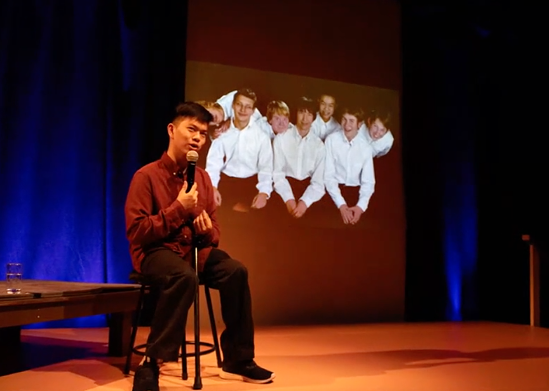 A man sits on stage with a microphone in front of a projector screen 