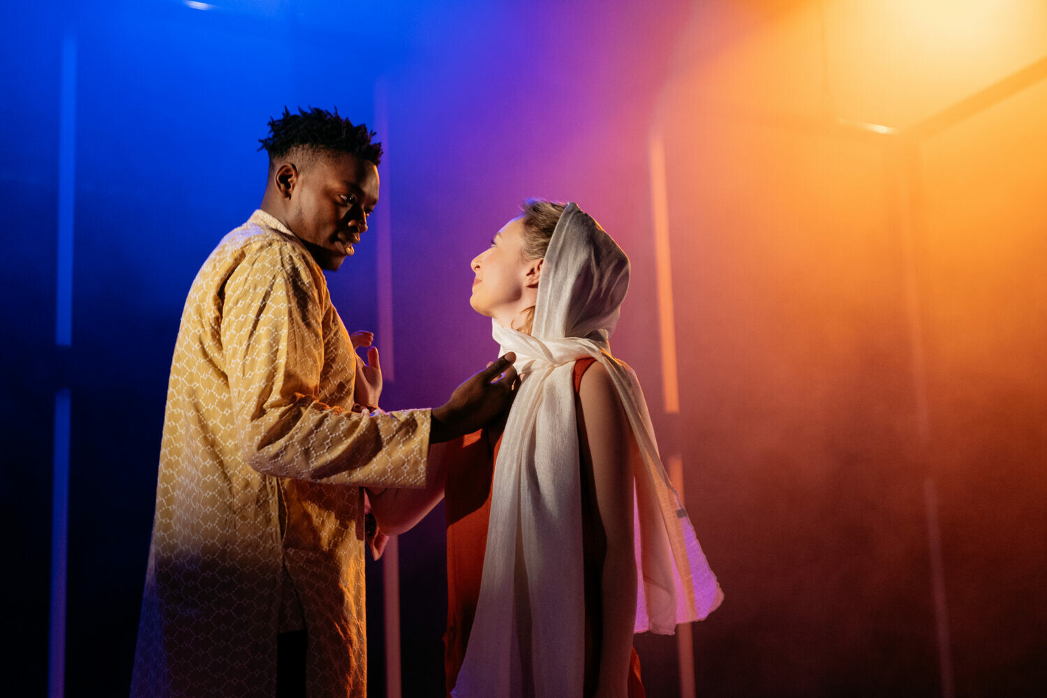 Two people stand on stage looking at one another in front of colorful lighting.