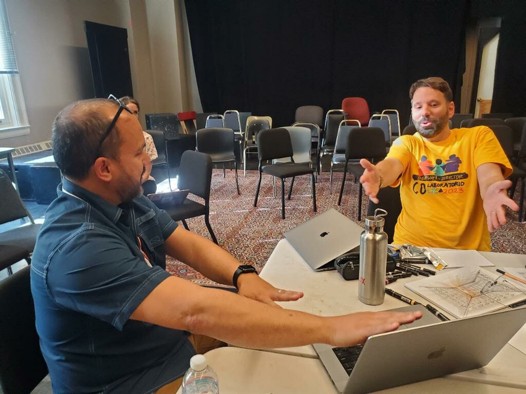 Two Artists sit at a table speaking at a workshop each with am open laptop in front of them.