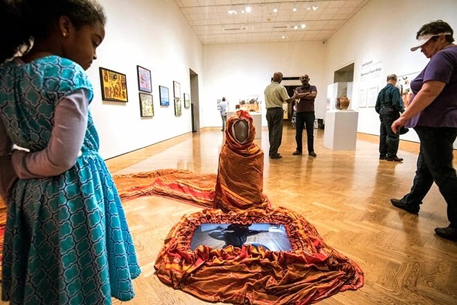 A figure wrapped in a blanket sits on the floor of a museum.