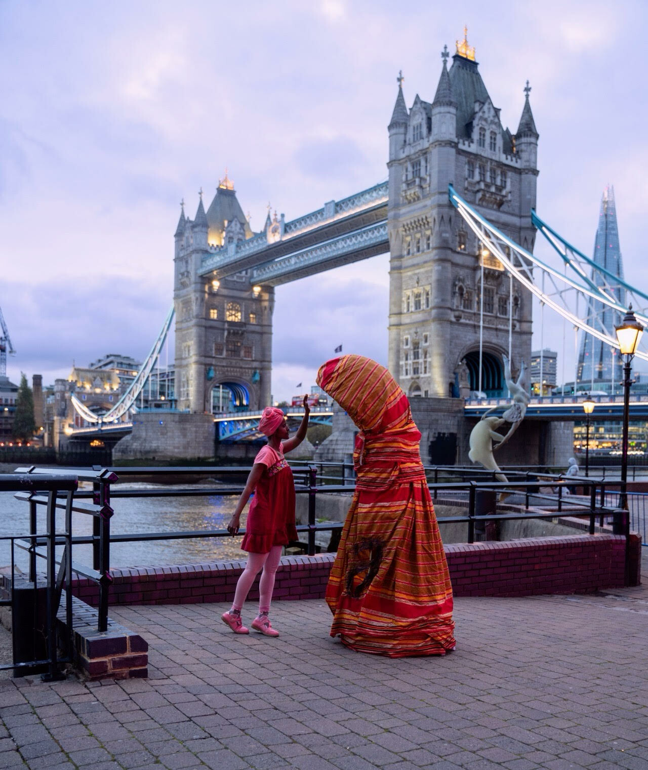 A woman shares space with a heavily costumed figure in red in front of a bridge.