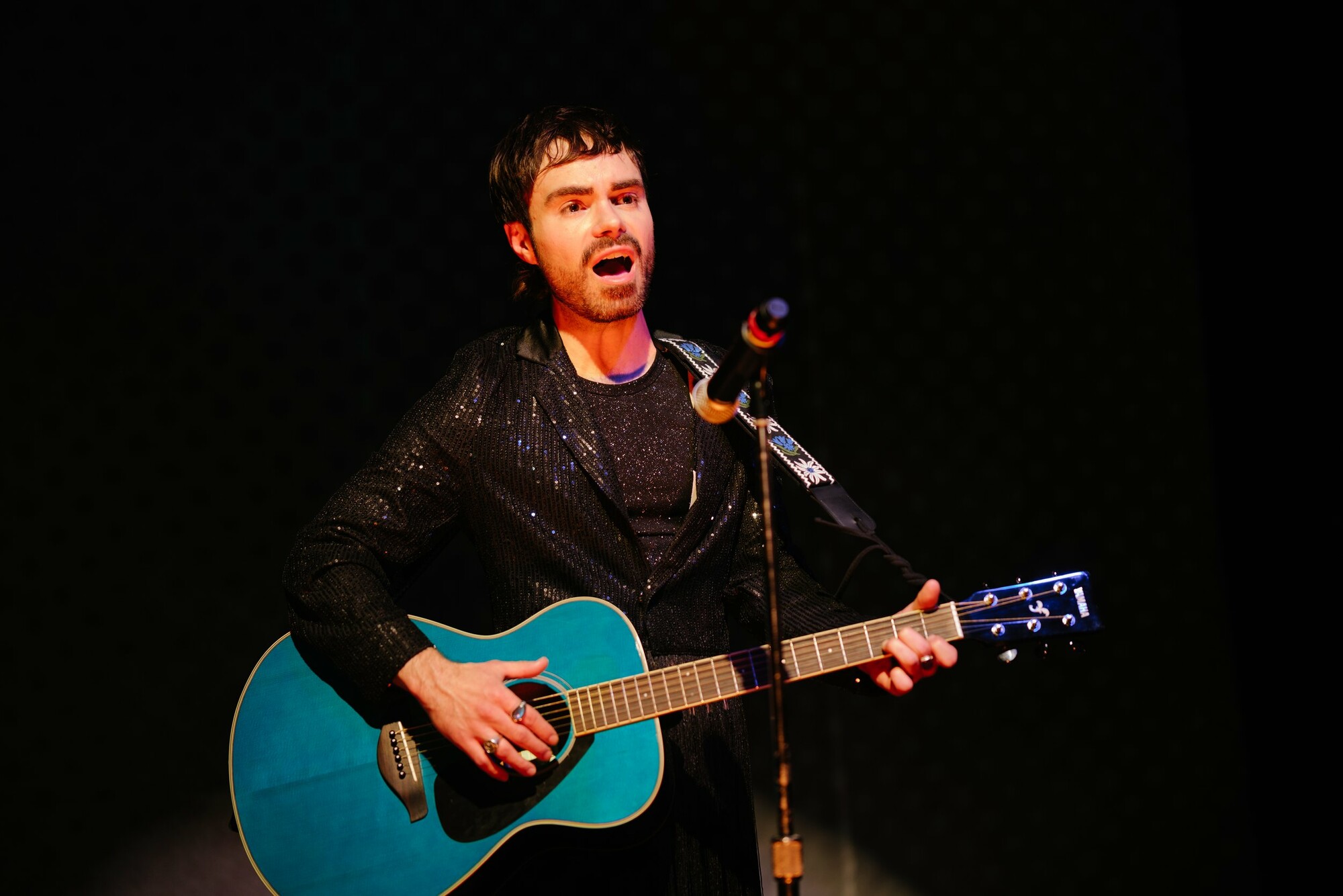 A man sings into a microphone while playing a blue acoustic guitar.