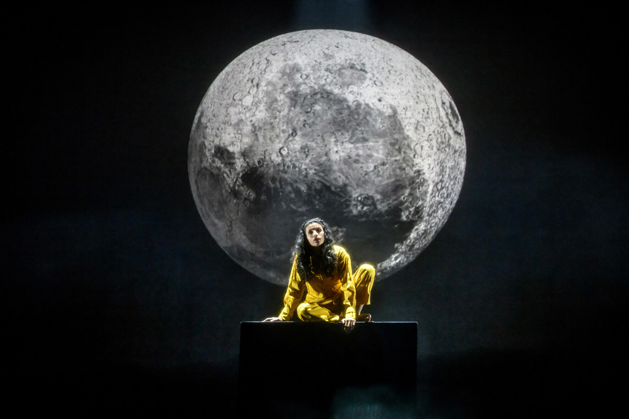 An actor sits on stage in front of a projection of the moon.