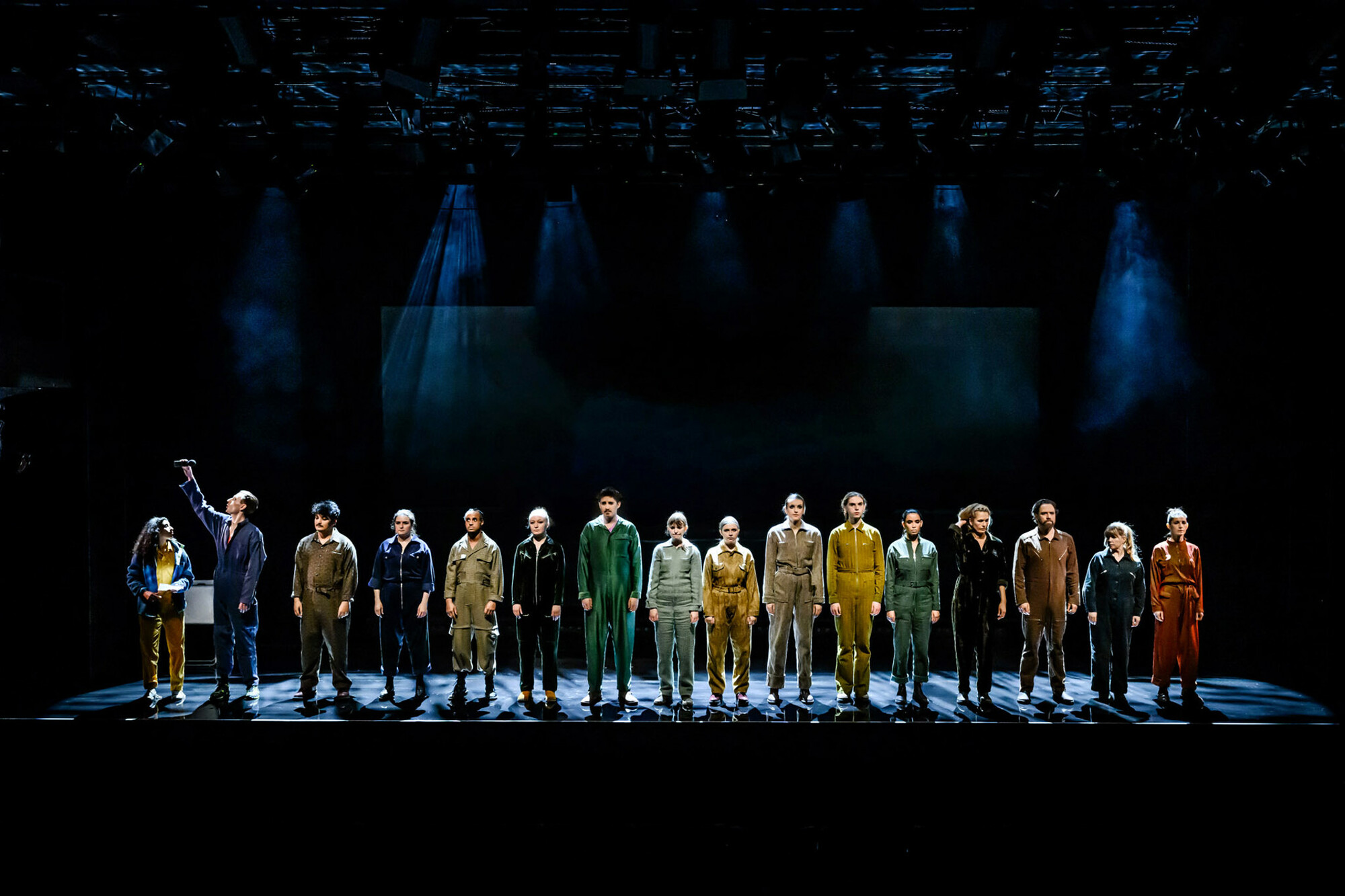 A group of actors stand in a  line on a dimly lit stage.