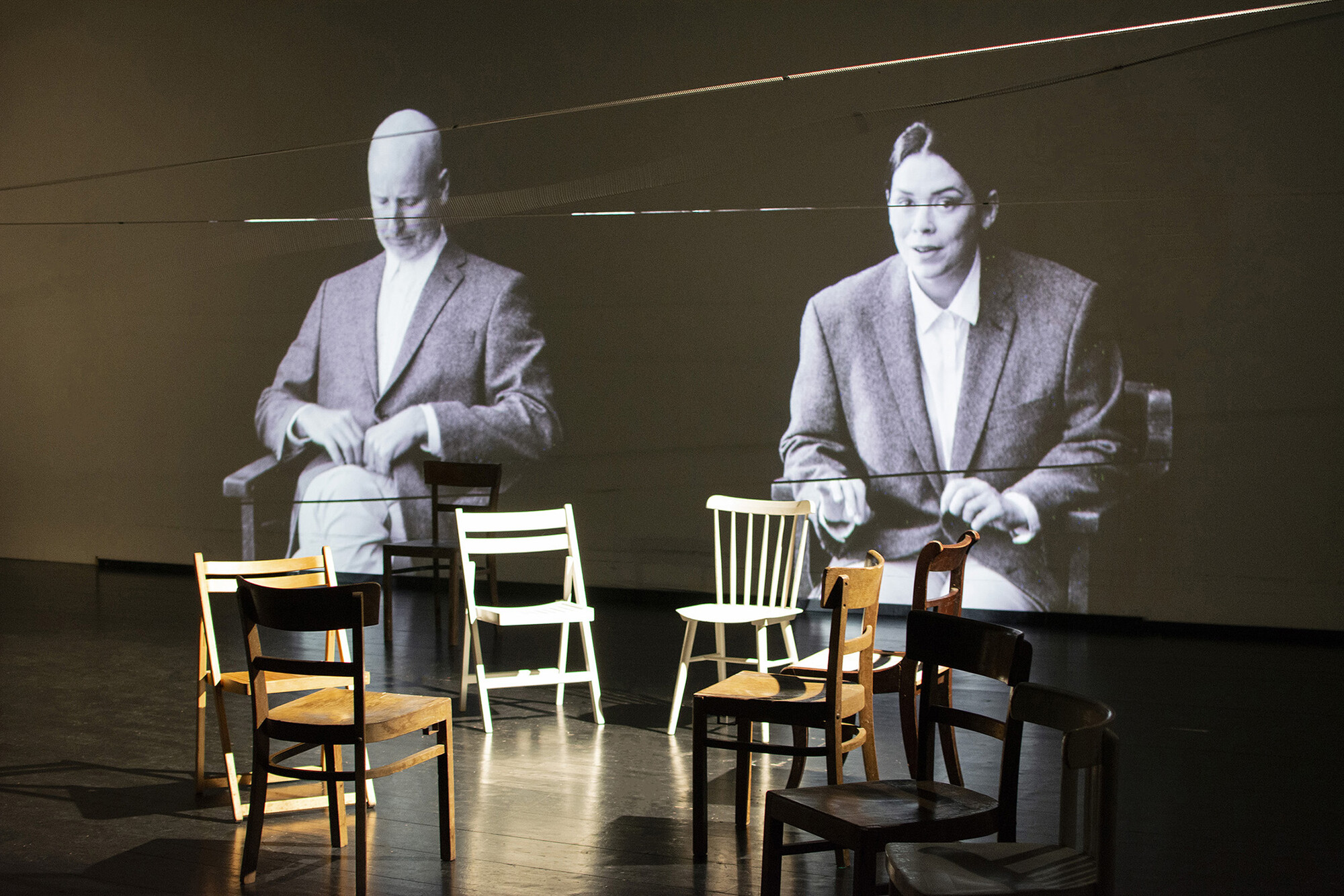 A projection of two actors behind a group of empty chairs on stage.